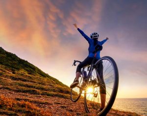 A woman celebrating on a bike on a hill next to the sea as the sun sets in the background