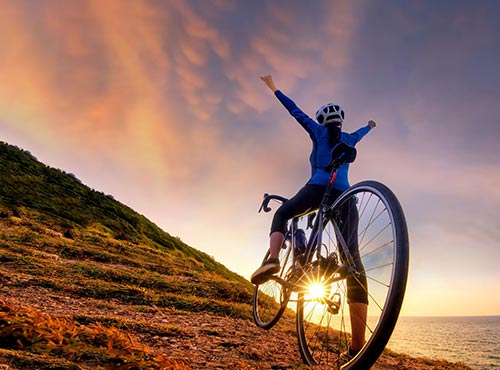 A woman celebrating on a bike on a hill next to the sea as the sun sets in the background